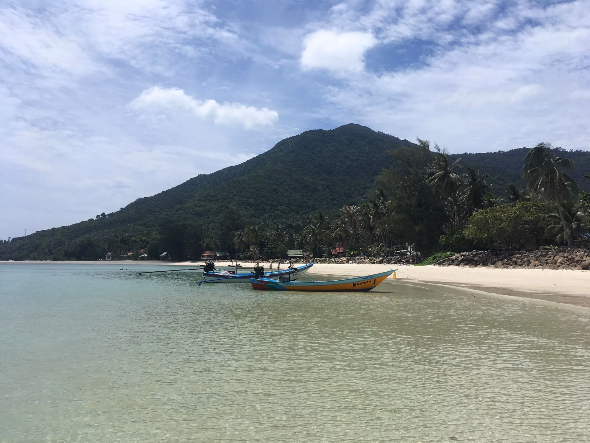 Clear, shallow water at Chaloklum beach, with longtail boats at anchor and a forested mountain rising behind the bay