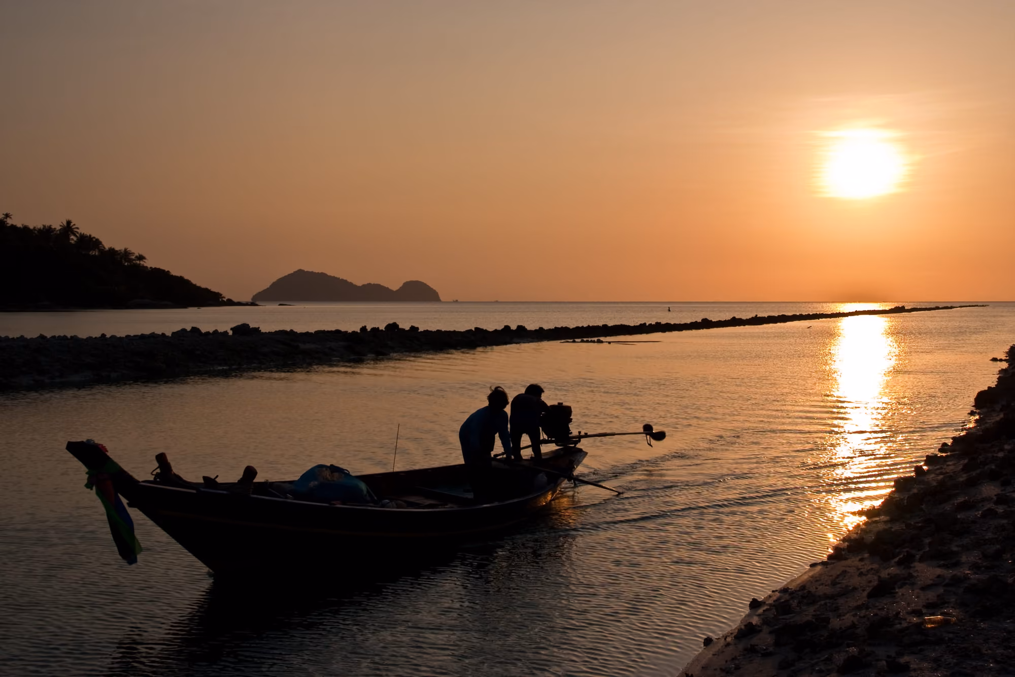 Two fishermen in silhouette push off a longtail boat at sunset in Ao Wok Tum, the sun reflecting on the bay