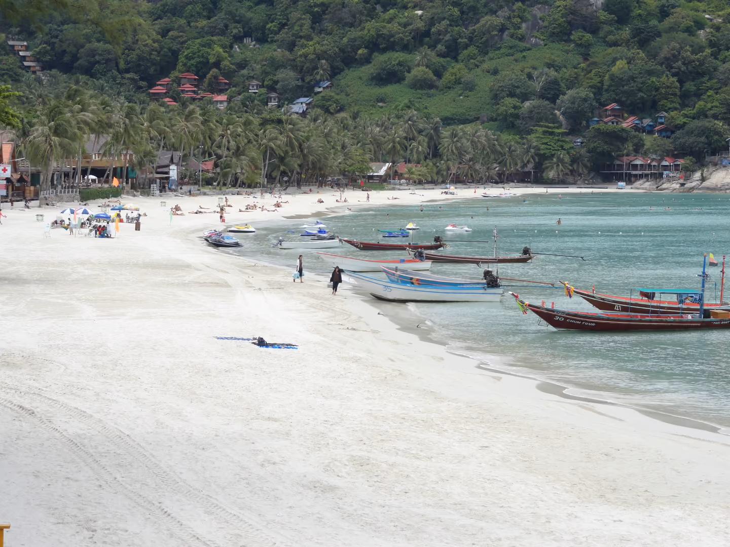 Wide white sand of Haad Rin Sunrise Beach in daylight, with longtail boats pulled up at the waterline and bungalows on the hill behind