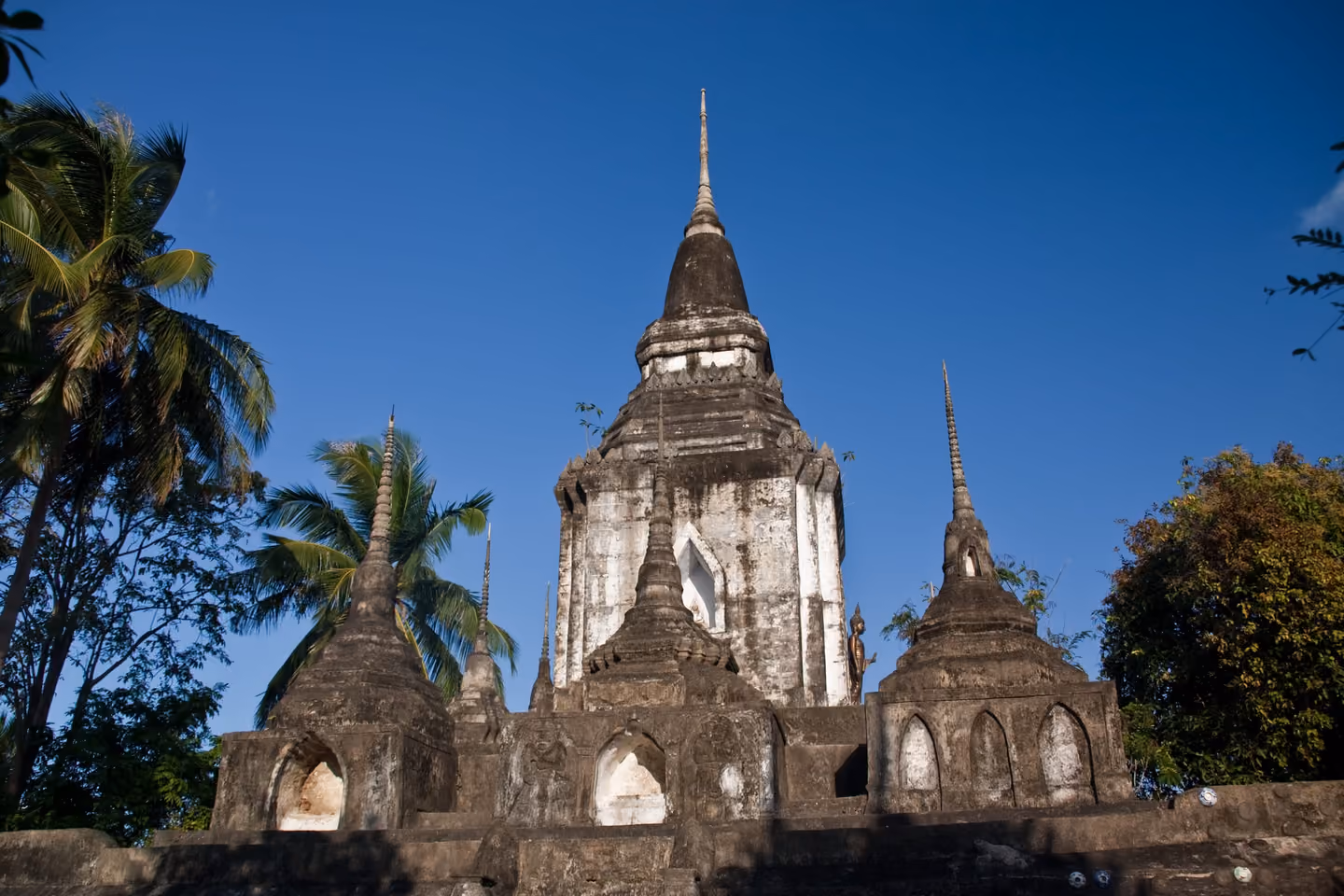 Weathered old chedis of Wat Phu Khao Noi, grey and white stone spires against a deep blue sky, with palms behind