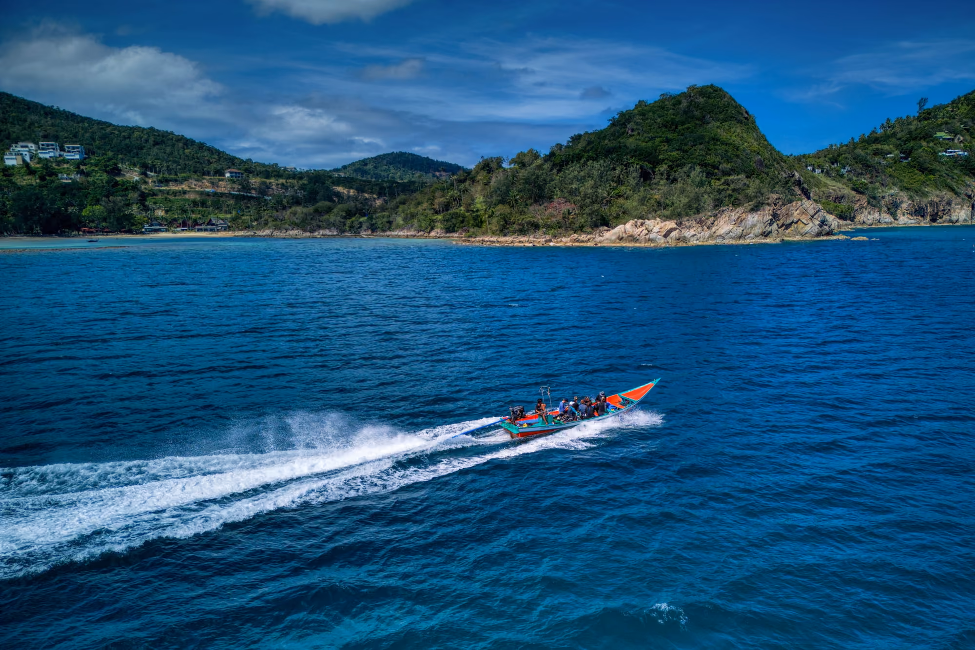 A longtail boat with passengers speeds across deep blue sea along a green, hilly coast, seen from above