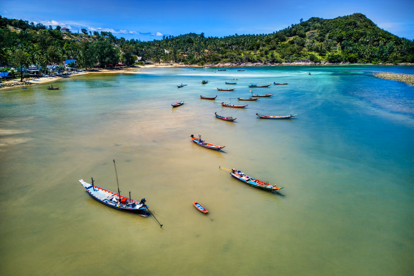 Aerial view of a shallow bay with about a dozen moored longtail fishing boats and a palm-lined shore below a wooded hill