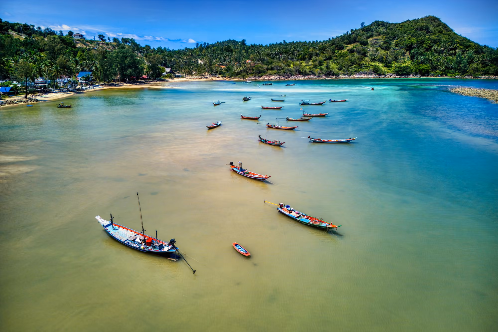Aerial view of a shallow bay with about a dozen moored longtail fishing boats and a palm-lined shore below a wooded hill
