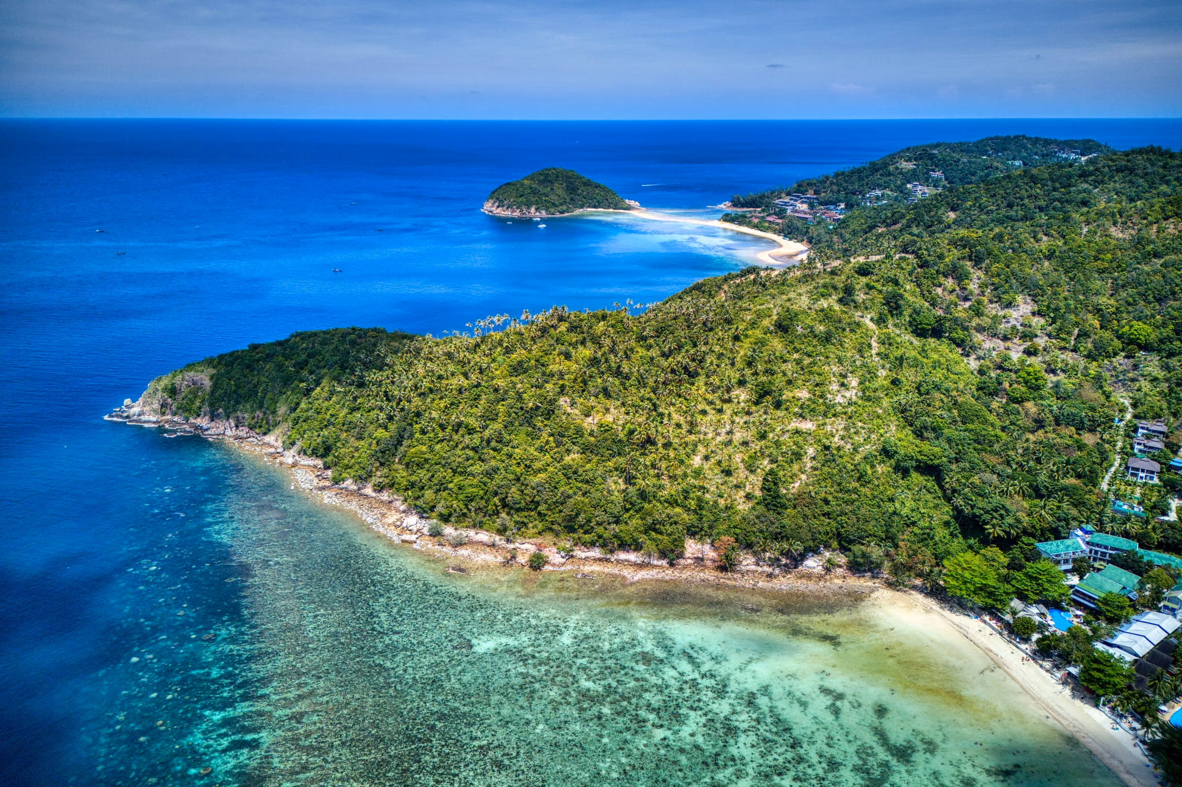 Aerial view of a forested headland on Koh Phangan with a coral reef in clear water and the islet of Ko Ma joined to the shore by a sandbar