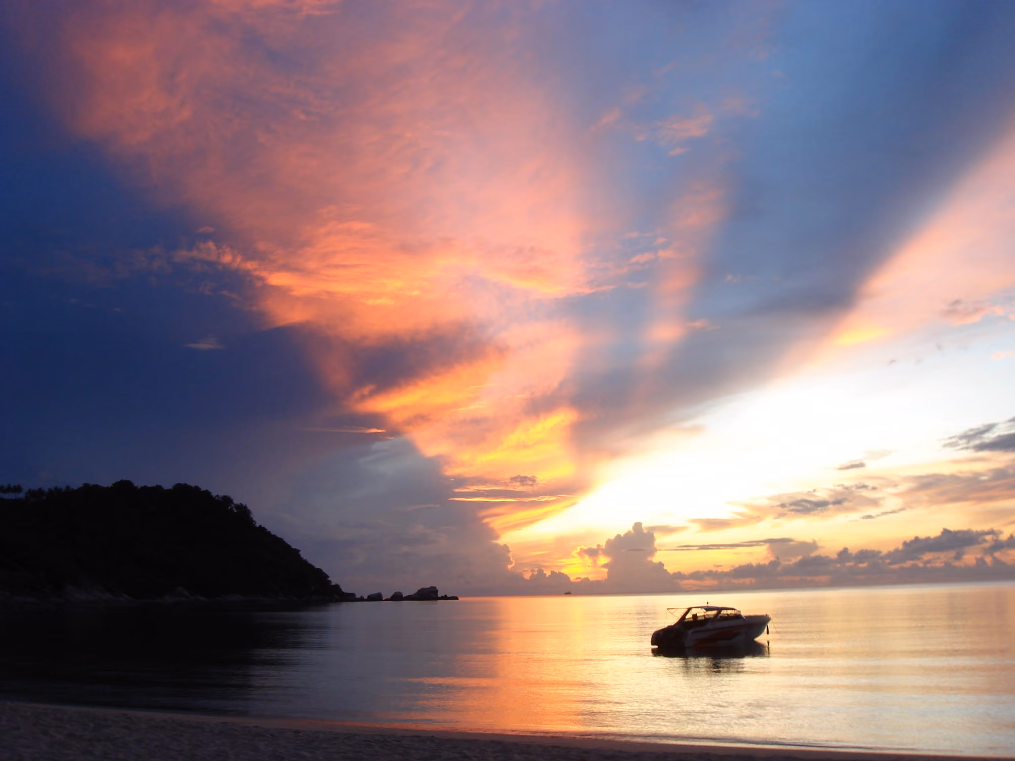 Sunrise over calm sea at Thong Nai Pan, with rays of orange light through tall clouds, a dark headland and a moored boat