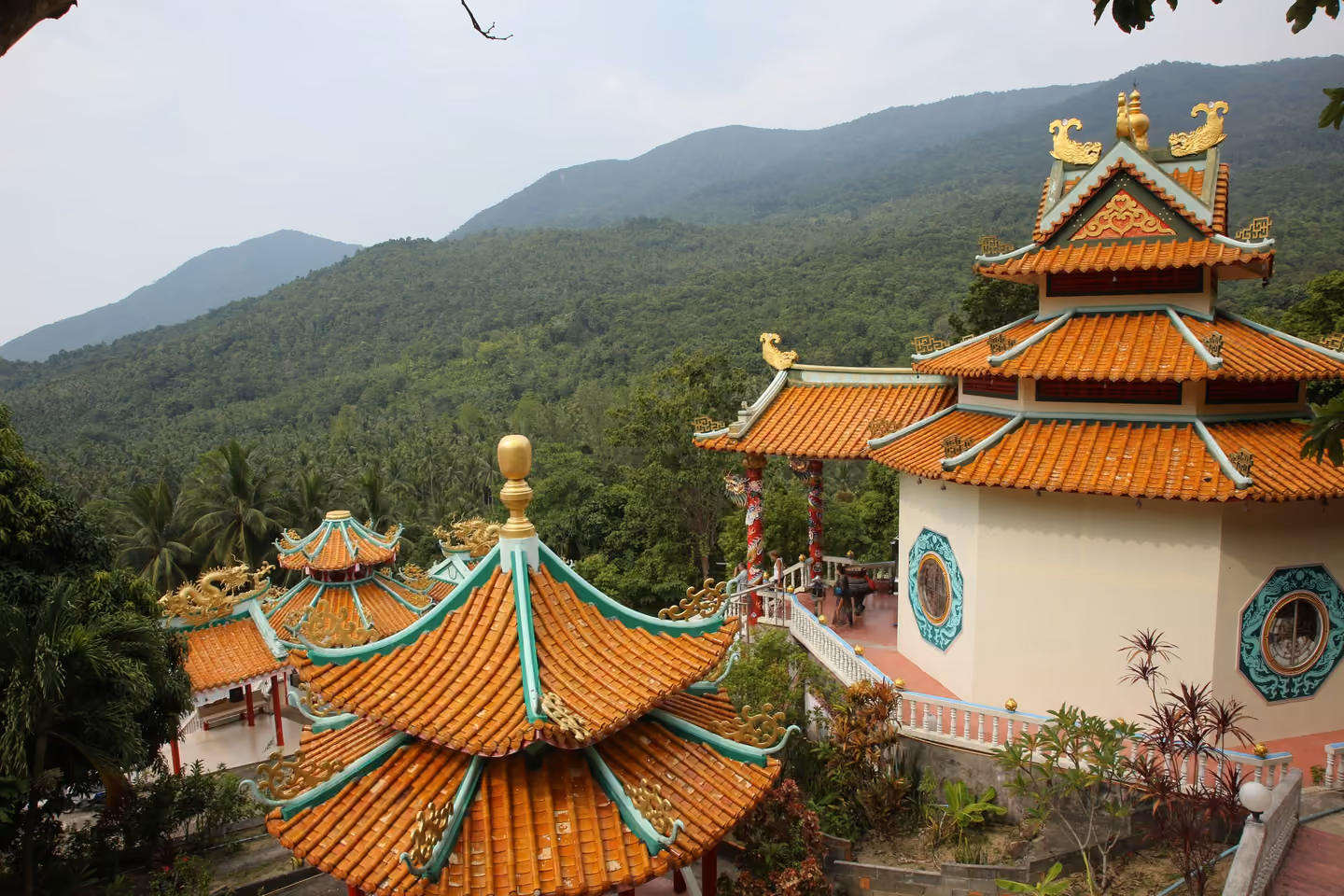 Orange tiled roofs with dragon ornaments at the Chao Mae Kuan Im shrine, above a valley of rainforest and hills