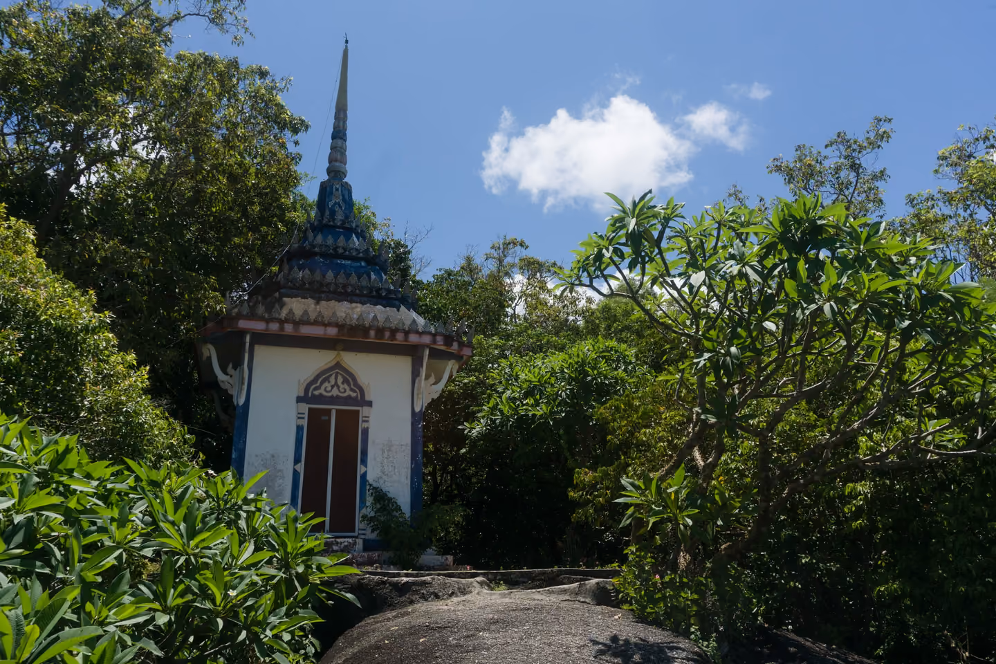 A small white shrine with a tall spire at Wat Khao Tham, half hidden by frangipani trees on a granite hilltop